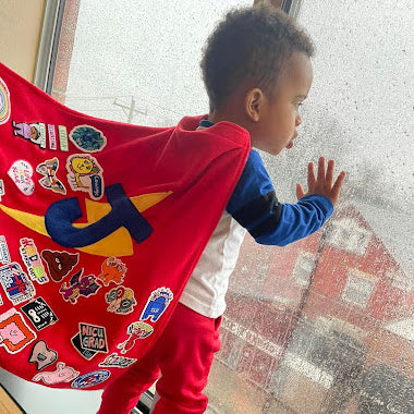 Photo of boy staring out of hospital window with his TinySuperheroes Cape on that is full of Patches.