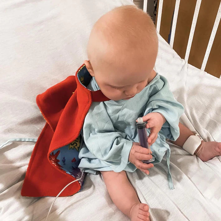Photo of baby boy in hospital gown and red TinySuperheroes Cape
